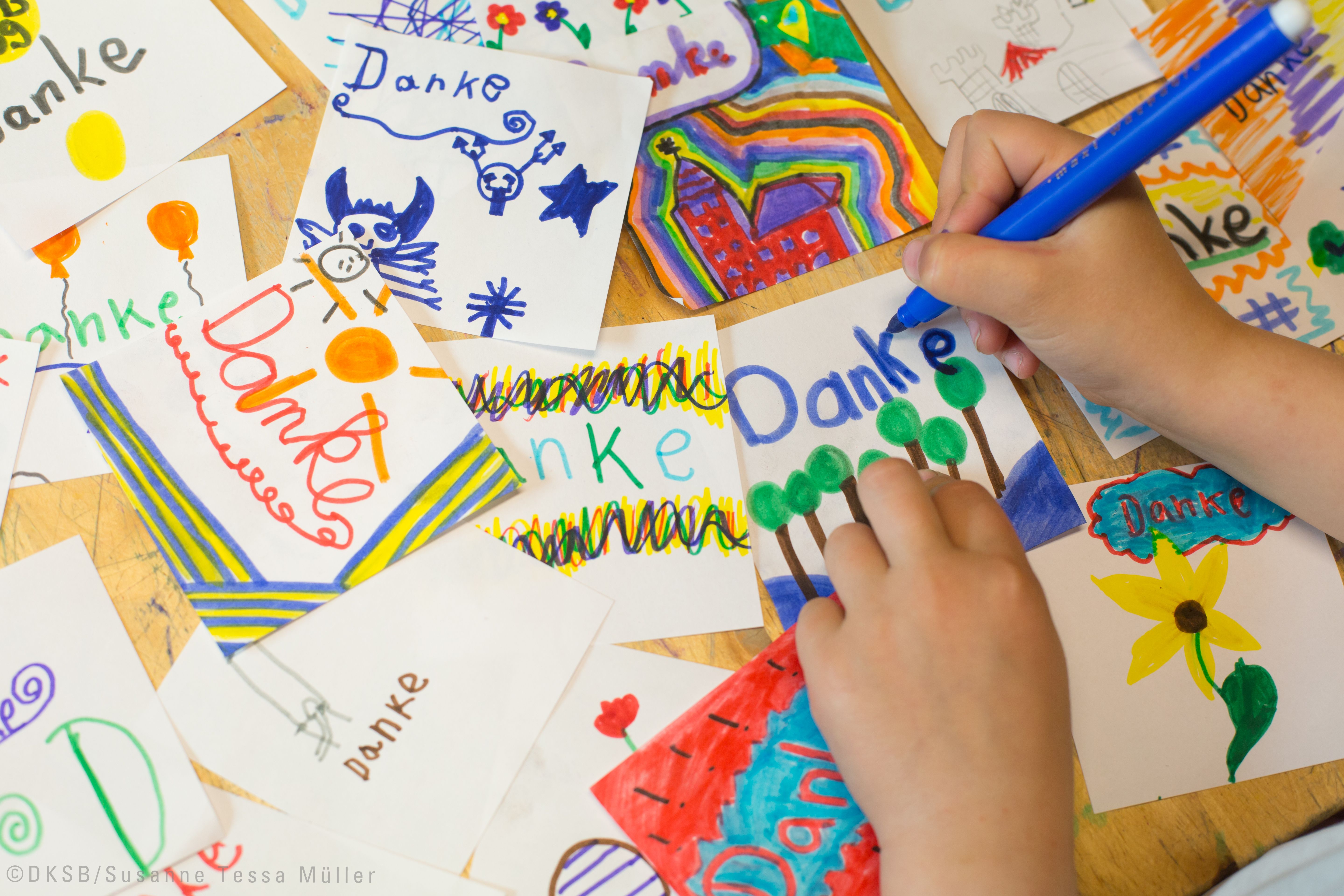 Ein Kind malt mit einem blauen Stift auf einer Karte mit dem Wort "Danke" umgeben von bunten Zeichnungen.