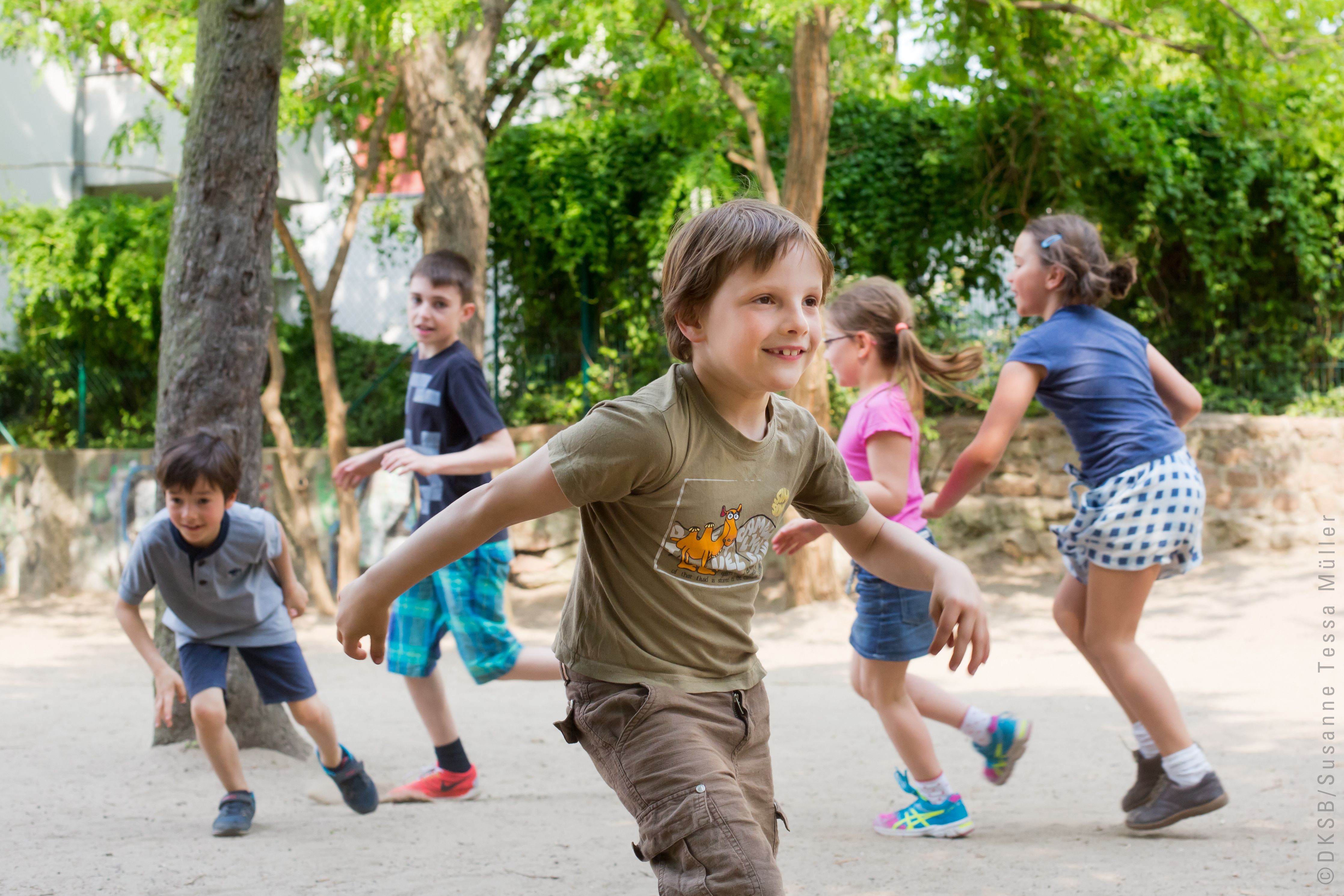 Kinder spielen fröhlich in einem Park, während sie miteinander rennen und lachen, umgeben von grünem Laub.