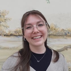 Eine lächelnde Frau mit Brille trägt einen beigen Cardigan vor einer malerischen Hintergrundlandschaft.